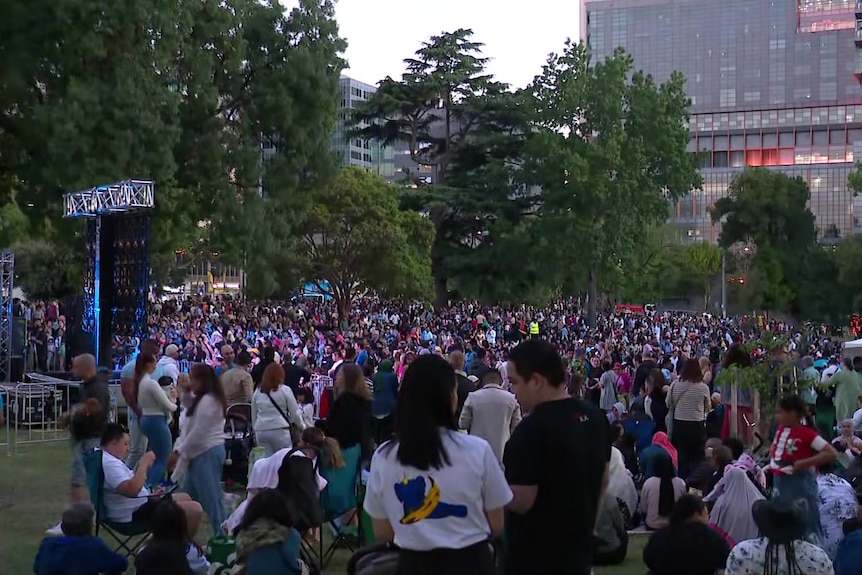 Large crowds cover the grass of a city park where a stage and some small tents have been set up, with highrises behind.