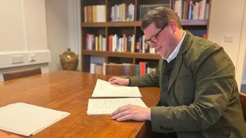 University of York An academic in a green jacket and glasses sits at a desk reading a pile of papers