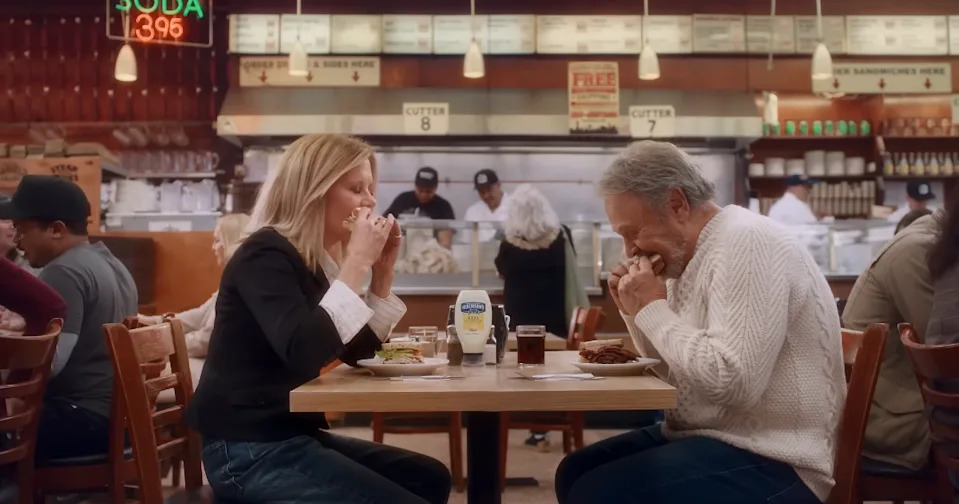 Two people sit at a deli table enjoying sandwiches, with a retro interior and deli counter in the background