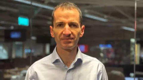 Dr Eoghan Ferrie is in a newsroom. The background is blurred. He's looking intot he camera. He has brown hair and is wearing a blue shirt. 