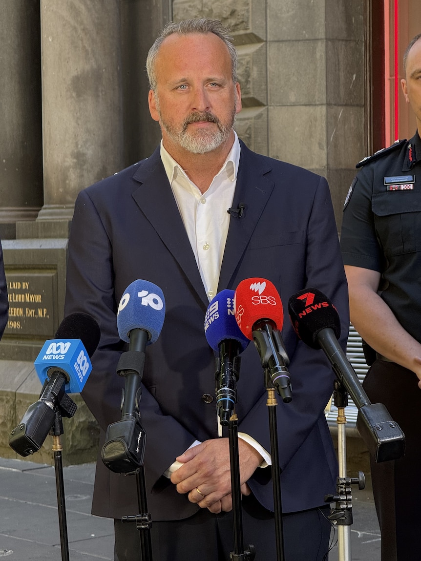 A man with grey hair, a moustache and beard in a navy jacket and white collared shirt stands behind microphones.