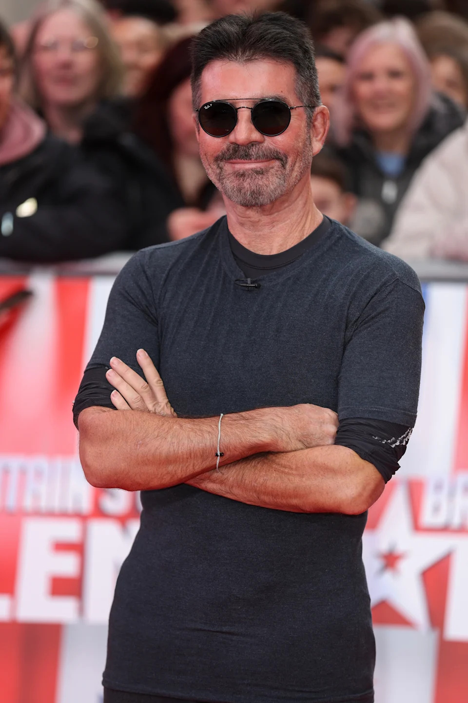 A man on the red carpet wears a fitted t-shirt, sunglasses, and has arms crossed, smiling at the event