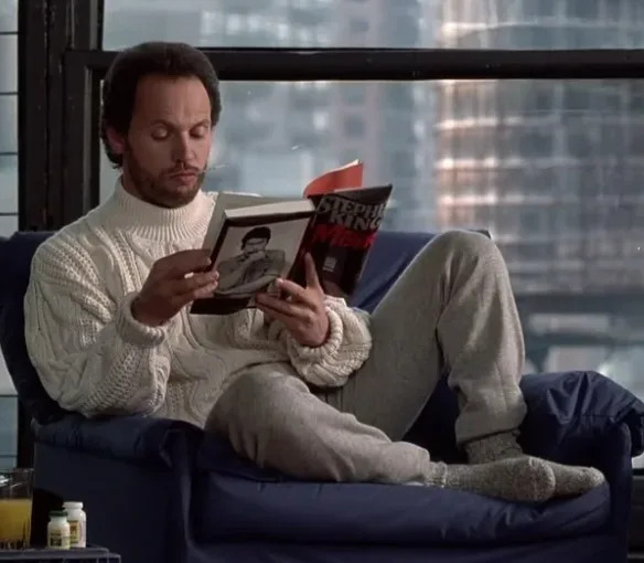 Person in a cozy setting reads Stephen King book, wearing a chunky knit sweater and sweatpants, with a cityscape visible through the window