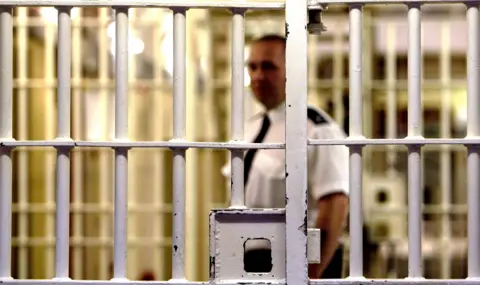 Getty Images A prison guard stands behind a locked white metal gate inside HMP Pentonville. The gate is in focus with the guard and background blurred. The guard is dark trousers, a white short-sleeved shift and a black tie.