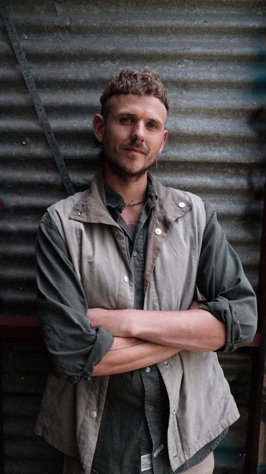 A man in neutral coloured clothes stands cross-armed in front of a corrugated iron wall.