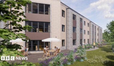 An artist's impression of the proposed care home which is a large brick building with large windows. There are brown wooden panels under the windows. There are two outdoor tables in front of the building to the left with white parasols. There are benches down the side of the building with people walking. To the right there is grass, trees and plants.