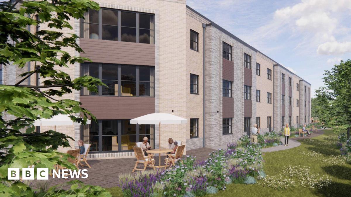 An artist's impression of the proposed care home which is a large brick building with large windows. There are brown wooden panels under the windows. There are two outdoor tables in front of the building to the left with white parasols. There are benches down the side of the building with people walking. To the right there is grass, trees and plants.