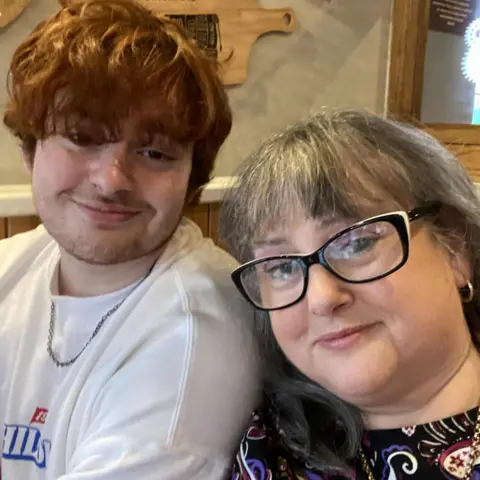 Maria Connolly A teenage boy with floppy ginger hair, wearing a white t shirt and silver chain. Next to him is a woman with long grey hair and black glasses wearing a patterned black top and gold chain. They ar eboth smiling and looking at the camera.
