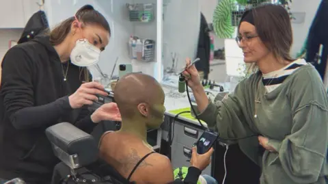 Olivia Jerrard Cynthia Erivo is sitting down in a black make-up chair in a movie trailer. She is scrolling on her phone while having her make-up done and is not facing the camera. Olivia Jerrard on the left is using a make-up brush near Erivo's ear and is dressed all in black while wearing a white face mask. On the right of Erivo is Alice Jones who is using an air brush machine to paint Erivo green.