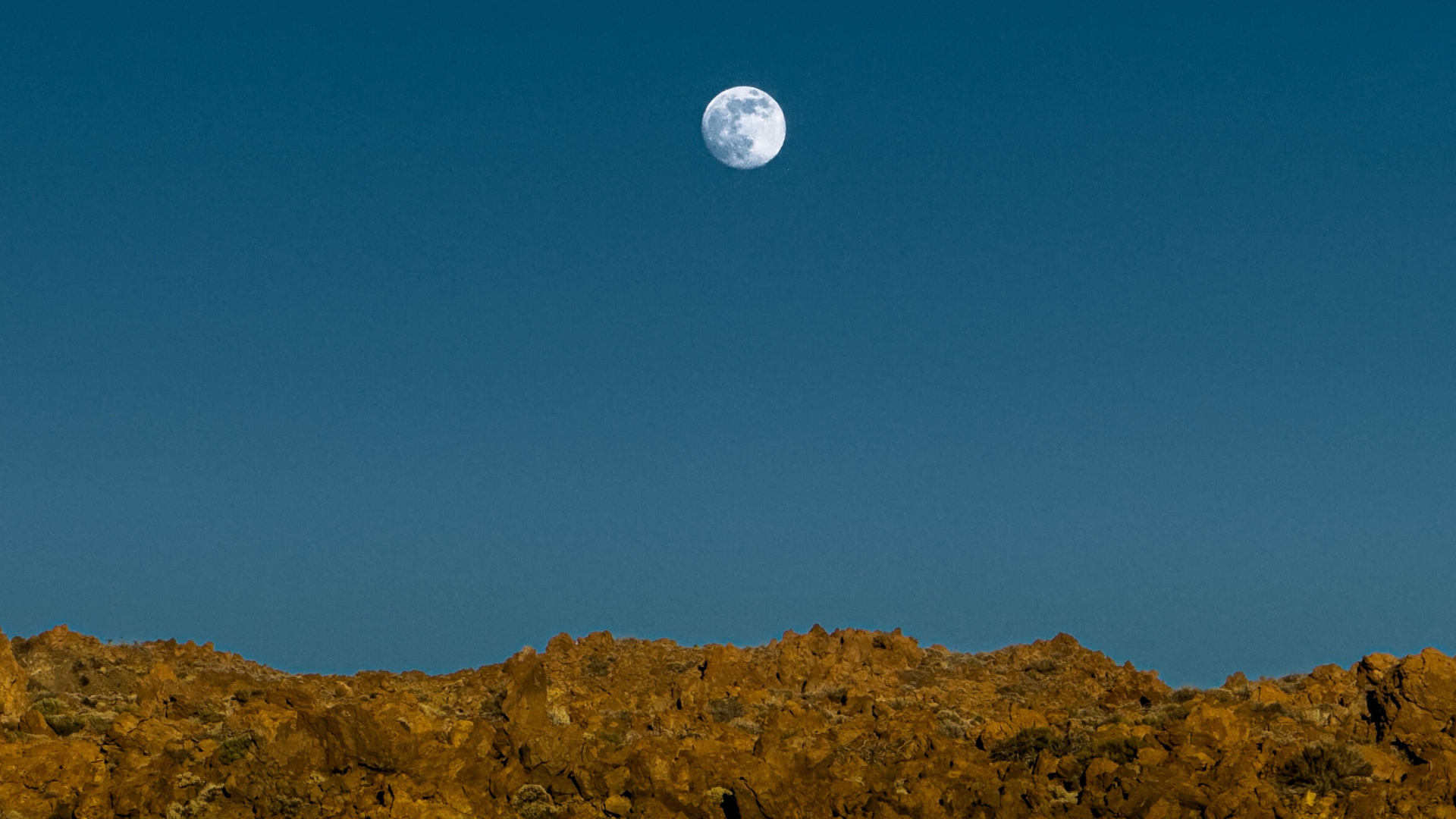 A full moon is pictured rising above a rocky landscape in a blue sky.