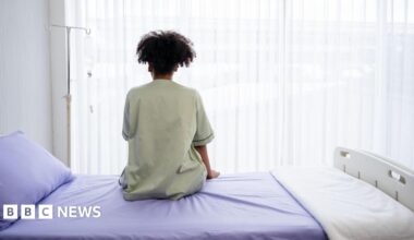 A person in a pale green hospital gown sits on a hsopital bed, made up with a lilac bedspread, pillow case and white blanket. They are facing away from the camera towards the window, with a gurney left of them.
