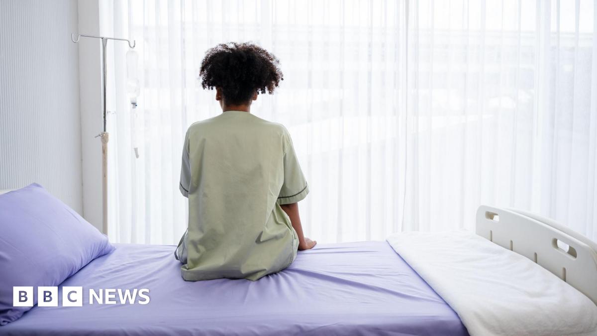A person in a pale green hospital gown sits on a hsopital bed, made up with a lilac bedspread, pillow case and white blanket. They are facing away from the camera towards the window, with a gurney left of them.