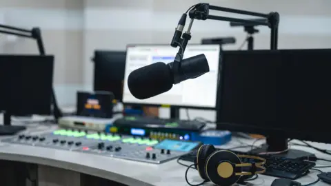 Getty Images A radio studio with a mixing desk and a microphone and a set of headphones on the desk. There are three computer screens with the one in the middle switched on.
