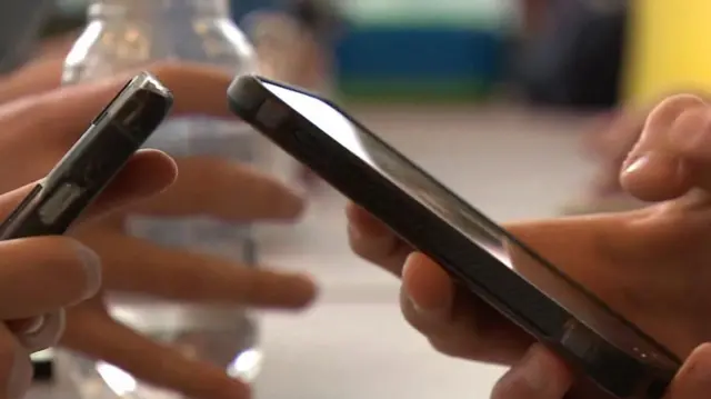 A close-up of hands holding smartphones, with one phone prominently in the centre being used. The setting appears to be a casual indoor environment, possibly a school cafeteria or common area, as there is a water bottle and blurred colourful objects in the background.