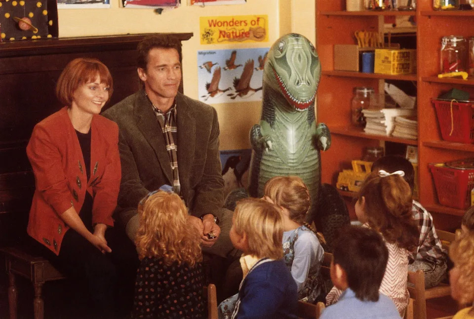 Two adults sit in front of a group of children in a classroom, next to a large dinosaur toy