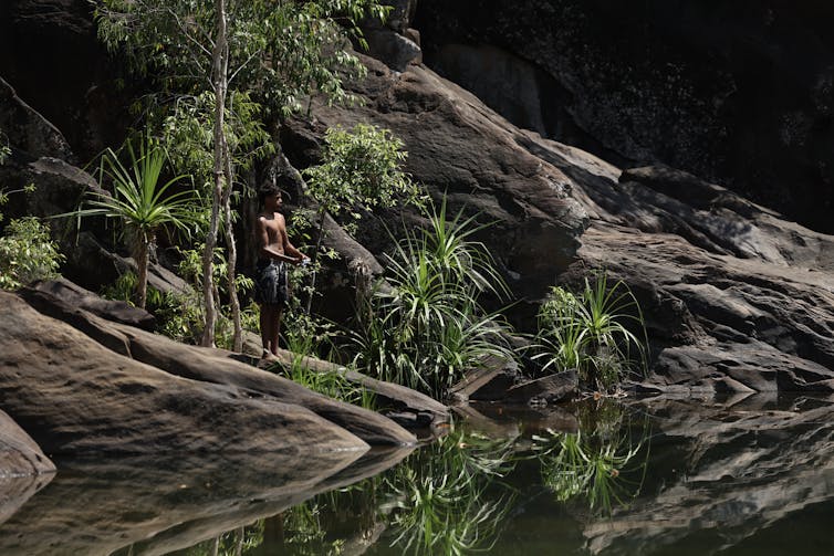 A young man holding a spear stands on the edge of a pool
