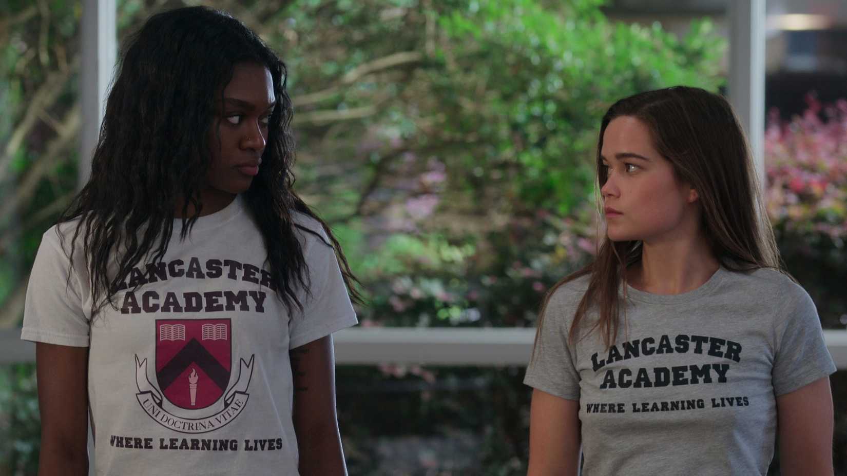 Calliope Burns (Imani Lewis) and Juliette Fairmont (Sarah Catherine Hook) glare at each other while wearing Lancaster Academy t-shirts in First Kill.