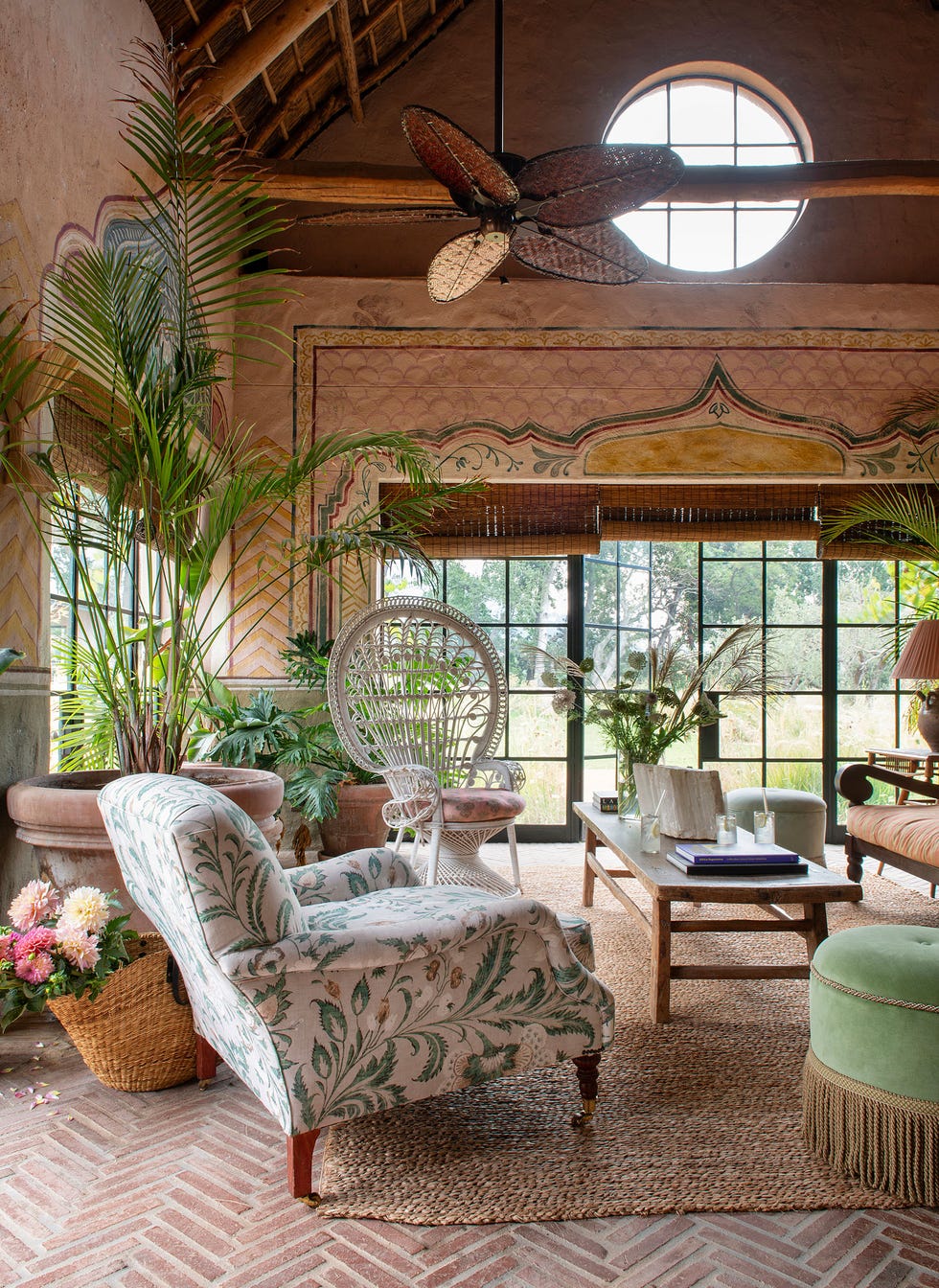 a living room with a terracotta chevron patterned floor and a wicker chair at the back and a large fauteuil in front with an ottoman in green velvet with fringe and large planters and patterned walls and glass doors at back
