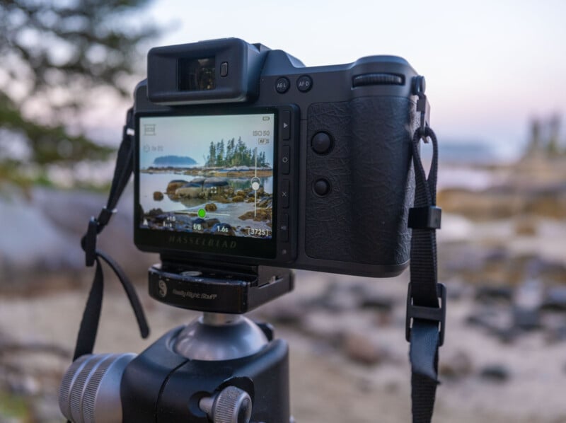A digital camera on a tripod, displaying a landscape scene with trees, rocks, and water on its screen, is set up outdoors near a shoreline with a soft, blurred background.