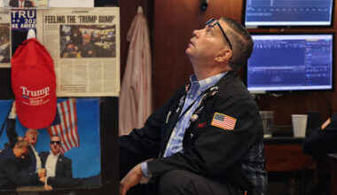 A trader on the New York Stock Exchange floor looks up, surrounded by images and headlines of Donald Trump and a red "Trump 2024" cap.