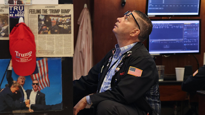 A trader on the New York Stock Exchange floor looks up, surrounded by images and headlines of Donald Trump and a red "Trump 2024" cap.