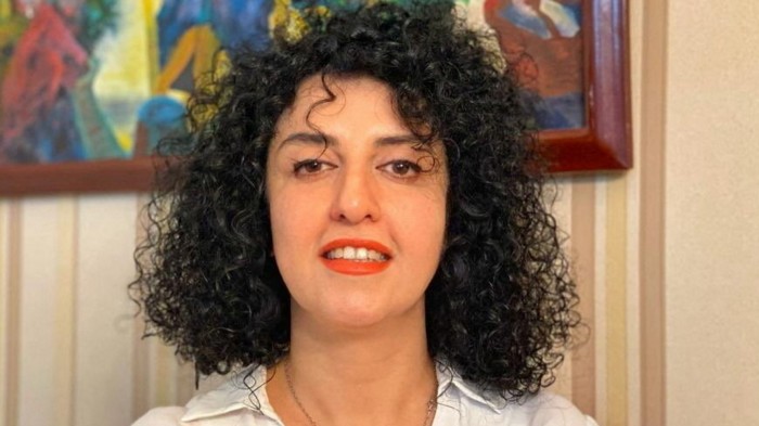 Narges Mohammadi poses indoors, wearing a white shirt, with curly hair and a colorful painting in the background.