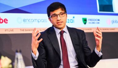 Nikhil Rathi speaks on stage, gesturing with both hands, wearing a suit and tie with a headset microphone.