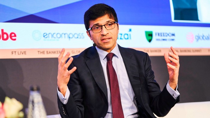 Nikhil Rathi speaks on stage, gesturing with both hands, wearing a suit and tie with a headset microphone.