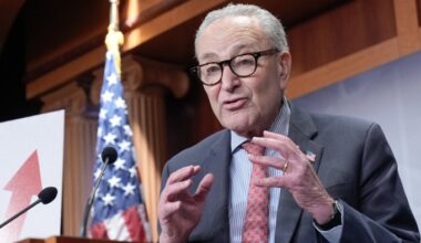 Chuck Schumer speaks at a podium with microphones, gesturing with his hands, and an upward arrow graphic and a US flag behind him.