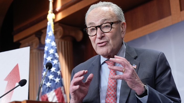 Chuck Schumer speaks at a podium with microphones, gesturing with his hands, and an upward arrow graphic and a US flag behind him.
