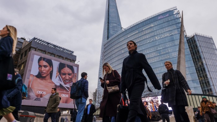 Commuters walk across London Bridge past a large ASOS advertisement, with The Shard and a modern glass office building in the background.