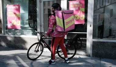 A Buyk delivery worker in branded gear stands with a bicycle outside a distribution centre in New York with Buyk advertising posters.