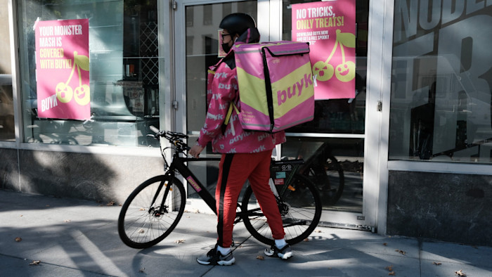 A Buyk delivery worker in branded gear stands with a bicycle outside a distribution centre in New York with Buyk advertising posters.