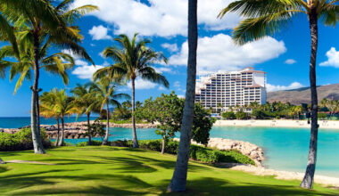 A white multi-story resort hotel stands near a sandy beach, surrounded by palm trees and turquoise water.