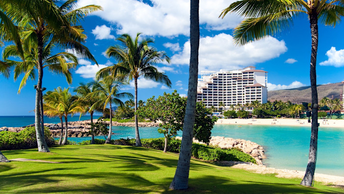 A white multi-story resort hotel stands near a sandy beach, surrounded by palm trees and turquoise water.