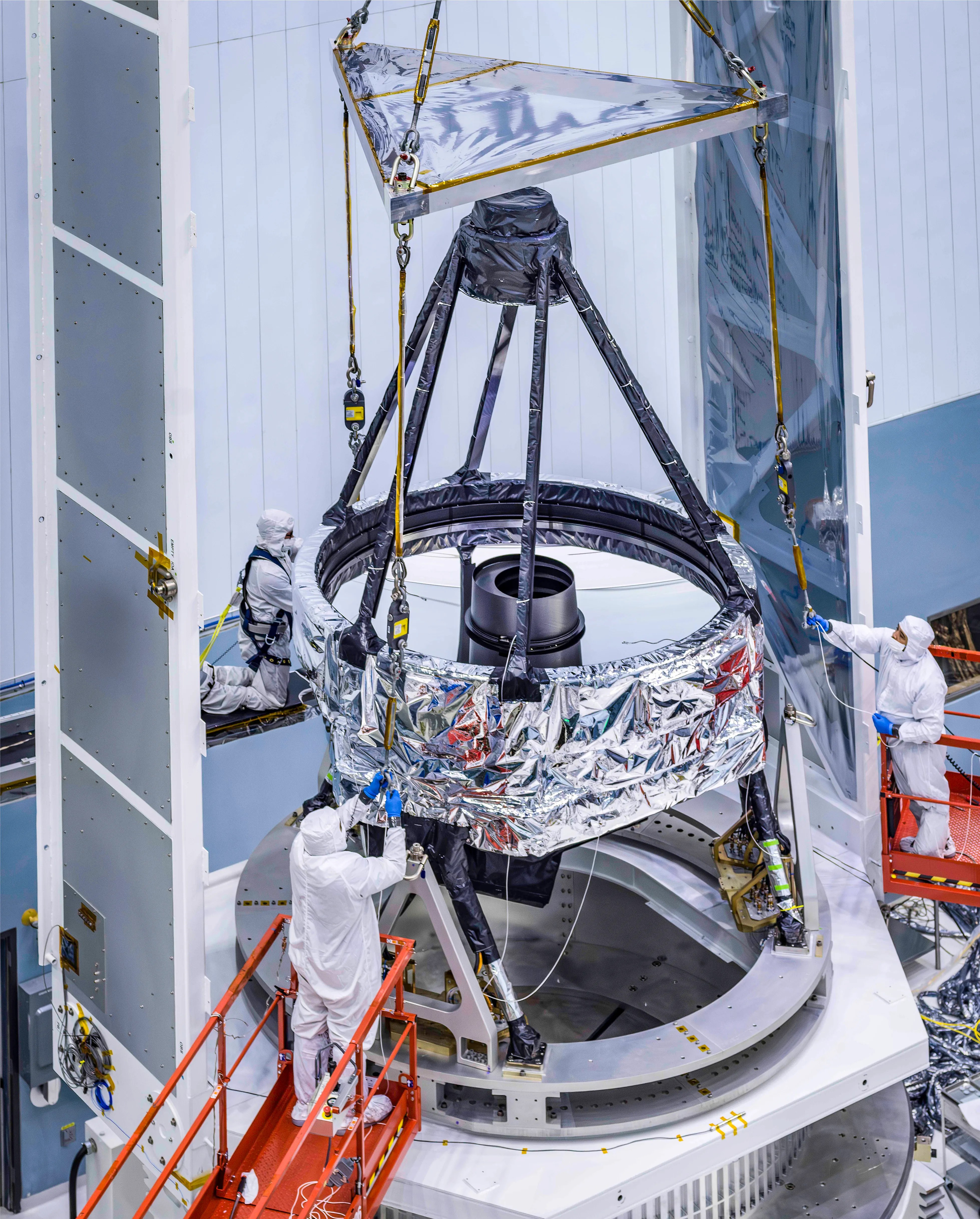 Technicians in clean suits work on assembling or inspecting a large, partially covered telescope component in a lab setting, using platforms and cranes—a testament to American science and innovation.
