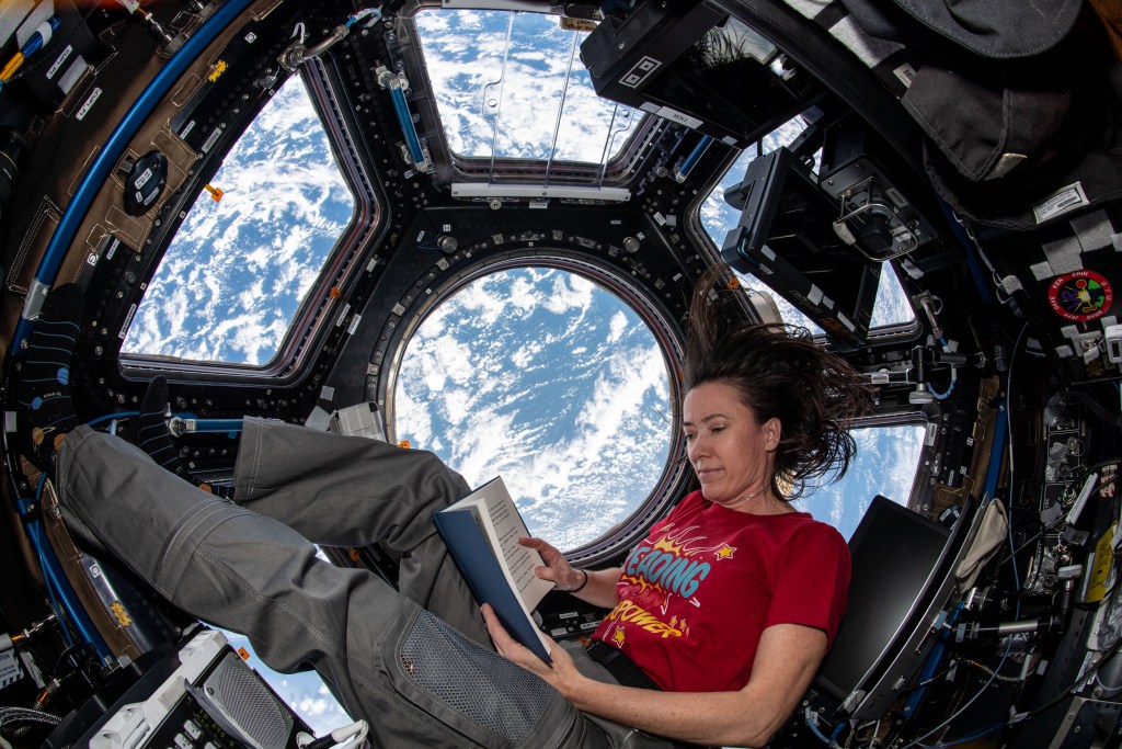 A woman with dark hair wearing a red shirt lounges with a book aboard the International Space Station. She is in the cupola--an array of windows that provide a breathtaking view of Earth below. Out the window, we see the Earth as mostly blue with white, streaky clouds.
