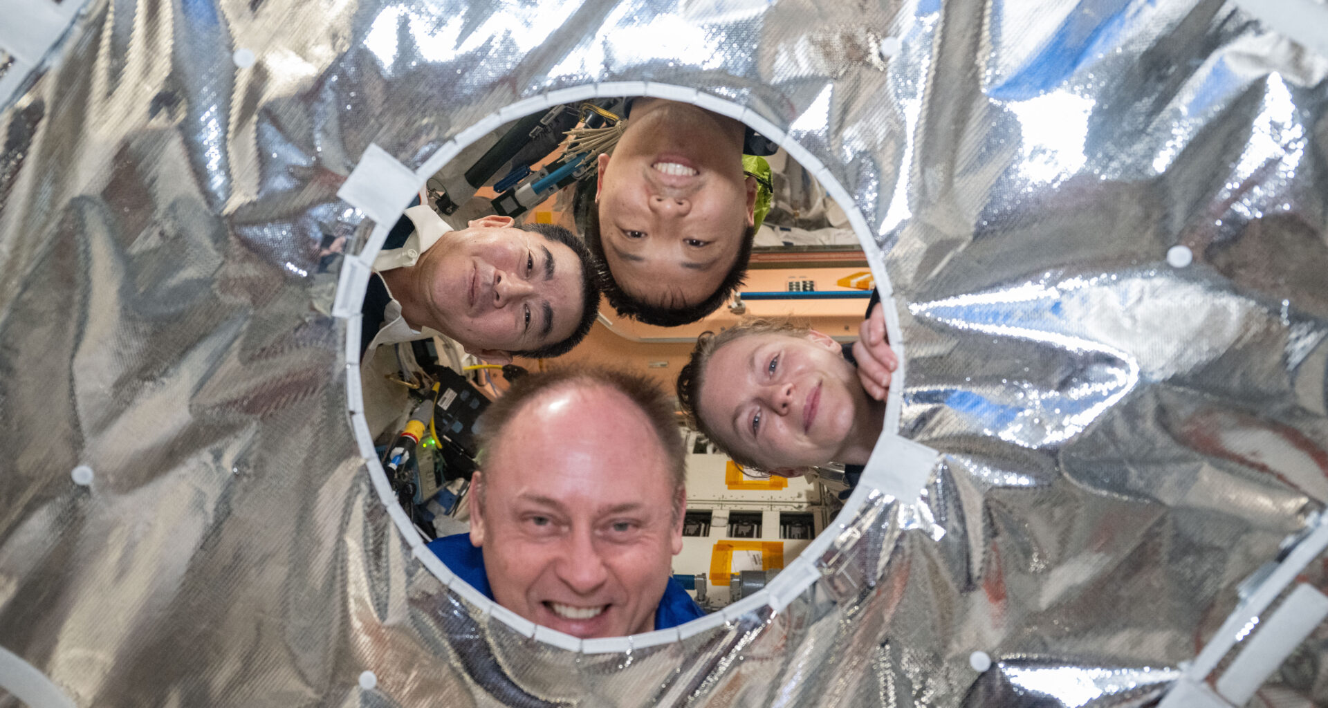Four people look through a circular hole in a silver metallic fabric. Clockwise from left are JAXA (Japan Aerospace Exploration Agency) astronaut Kimiya Yui and NASA astronauts Jonny Kim, Zena Cardman, and Mike Fincke.