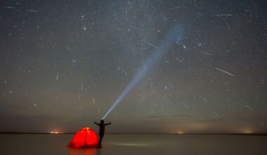 A person stands with arms outstretched behind a bright orange tent looking up at the night sky full of streaks with meteors with a large flashlight beam pointed up.