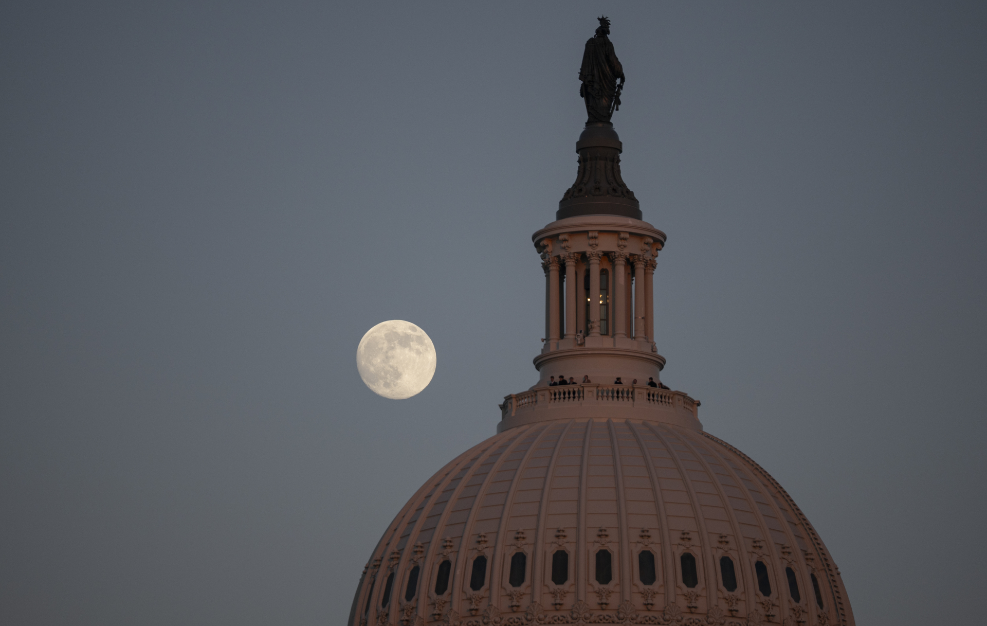 The full moon shines to the left of the dome of the Capitol Building in Washington D.C. in a darkening evening sky.
