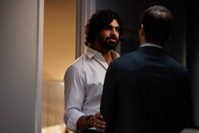 A curly-haired man in a white shirt stands in a doorway, resting a hand on the arm of a man in a dark suit whose back is to the camera, as they talk in a dimly lit hallway.