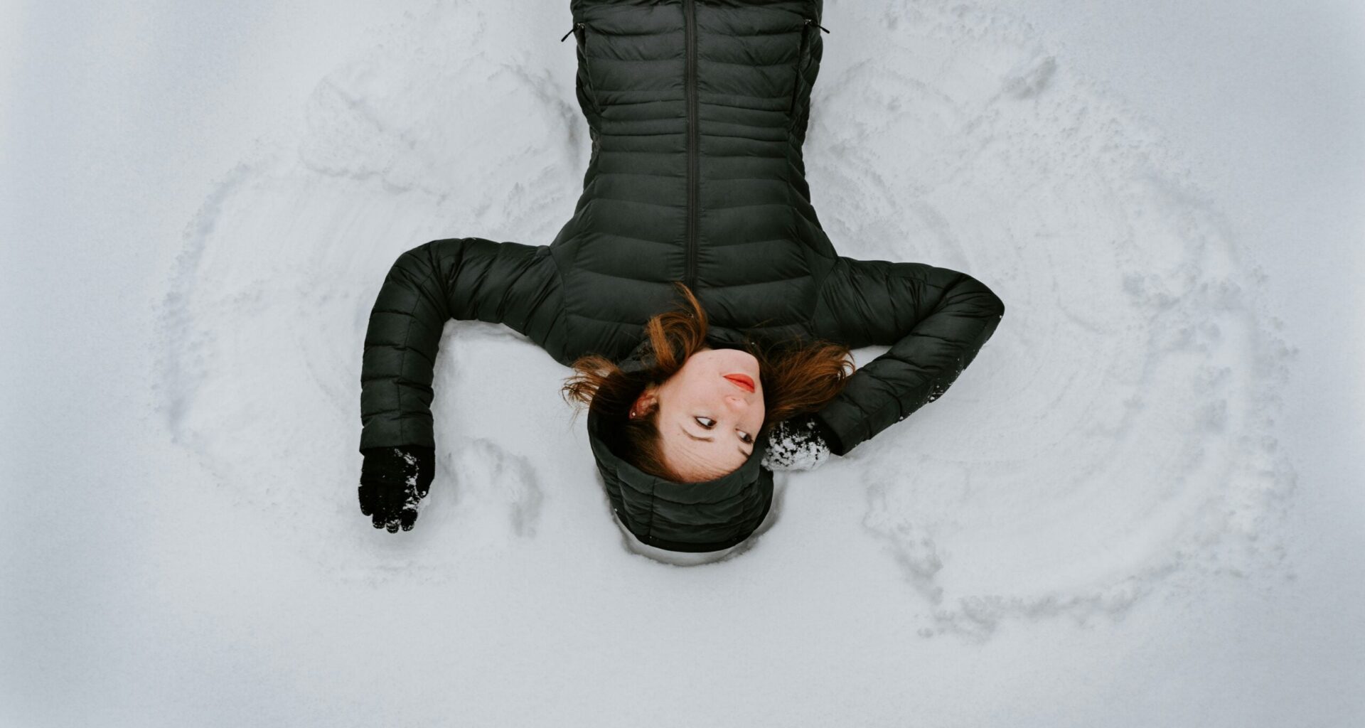 Woman in black winter coat making snow angels in snow.