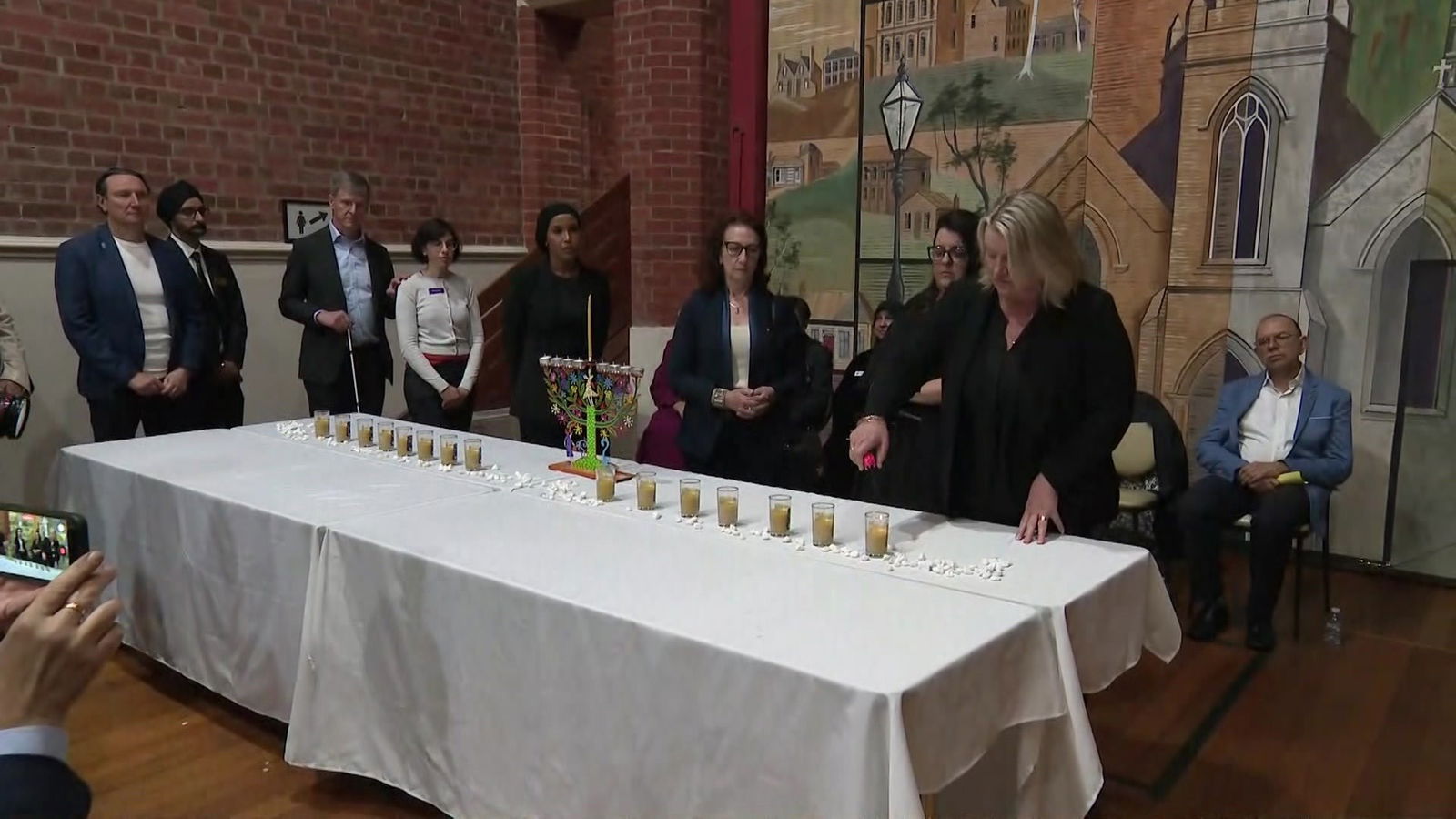 A woman with blonde hair dressed in black lights the first of a line of candles as people watch.