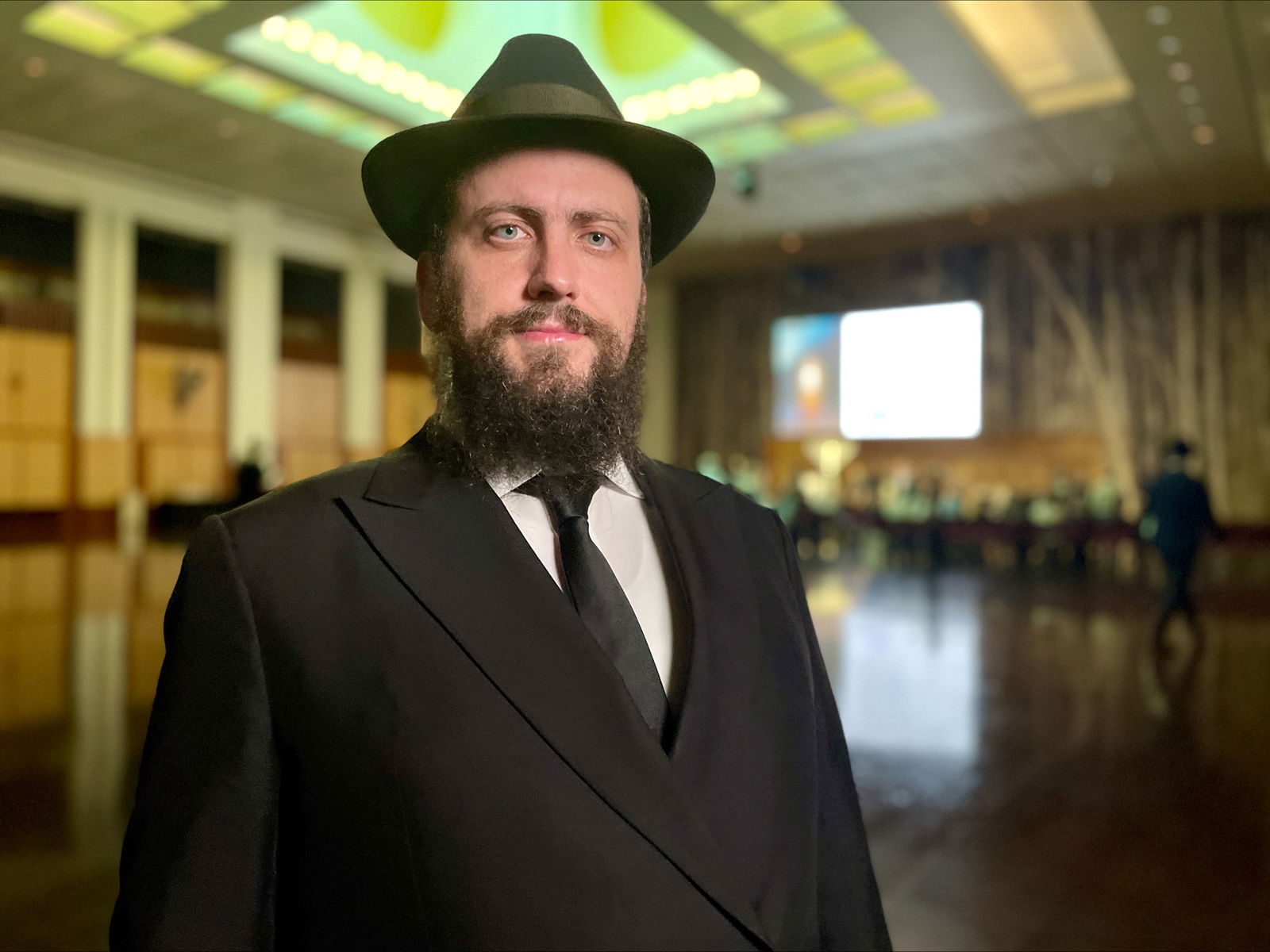 A man with a dark beard in a black hat, black suit, white shirt and black tie stands inside a hall.