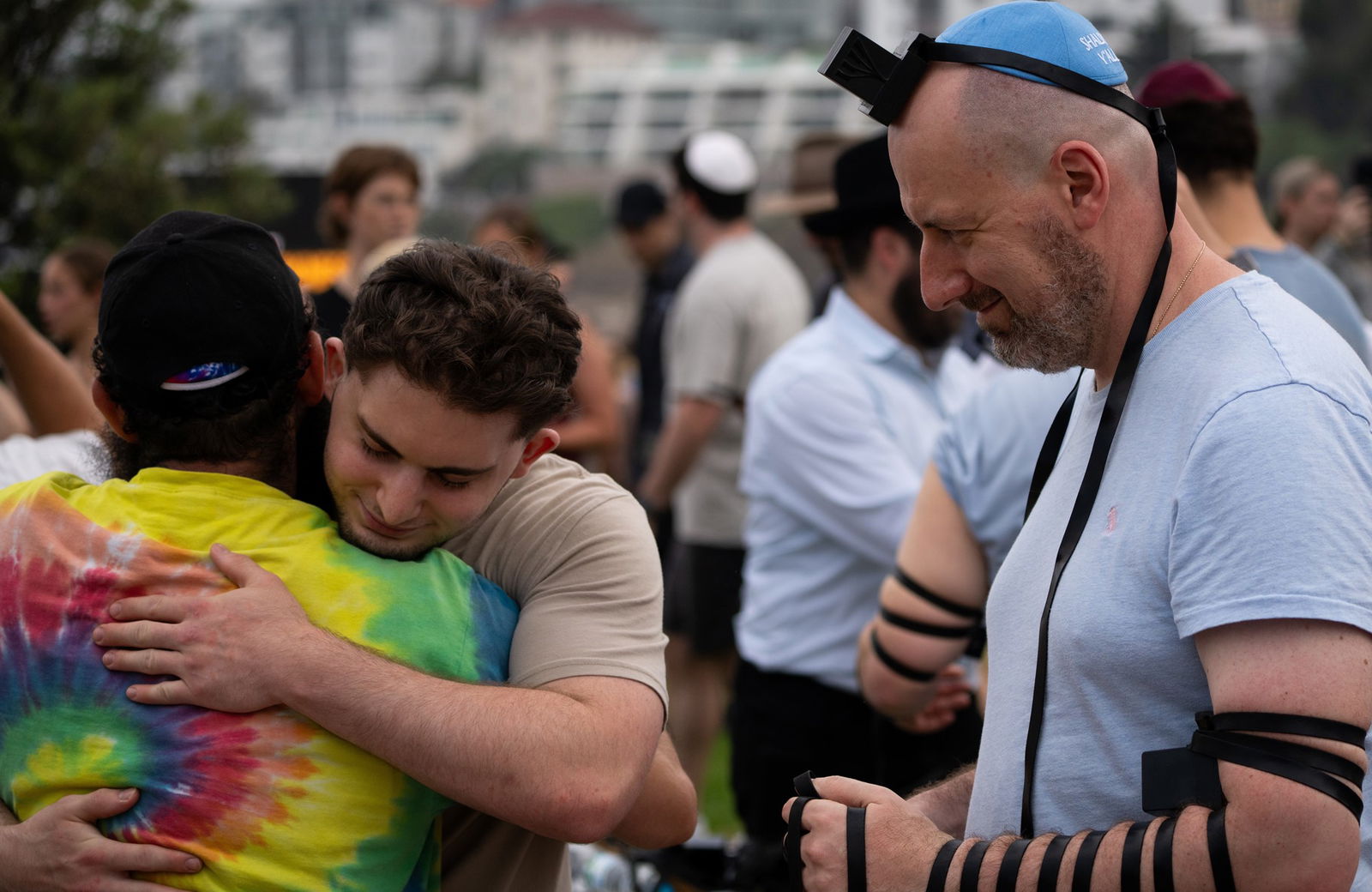 Two men embrace as another man, with a black leather strap wrapped around his arm, looks on smiling.