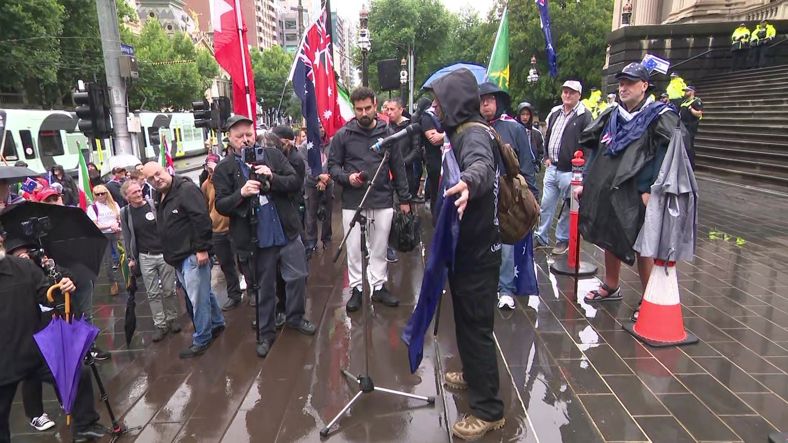 A man speaks into a microphone surrounded by people, some who have an Australian flag around their shoulders.