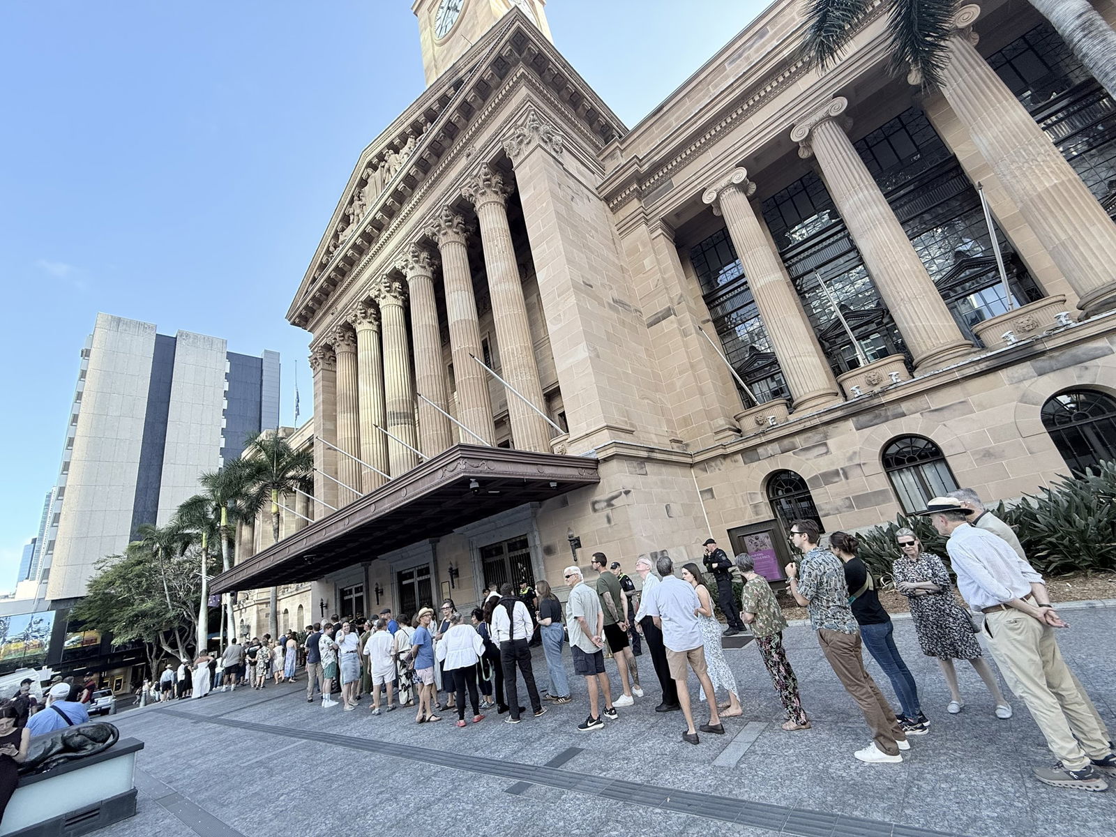 A long line of people stand in a queue outside an historical sandstone building under a blue sky.