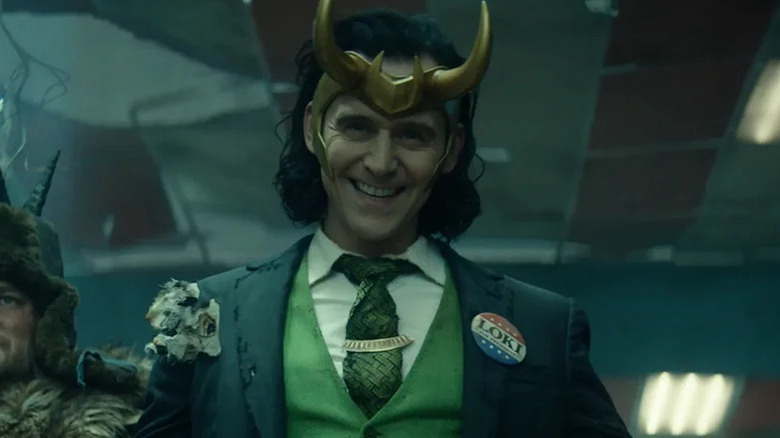 Loki (Tom Hiddleston) grins while wearing his horned headpiece and a three-piece suit with a campaign button in Loki