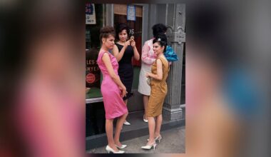 Four women in colorful, form-fitting 1960s dresses and white heels stand on a city sidewalk near a storefront, chatting and posing. Two are facing the camera, while two are turned slightly away.