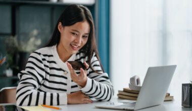 A young woman sitting with a laptop in front of her, talking to the smartphone, smiling.